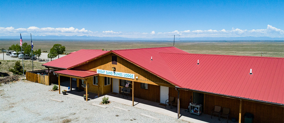 Exterior view of Lodge with signage over the door. Strands of clouds fill the background sky contrasted by the dark hues of mountains, and the gradient blues of the open sky. 