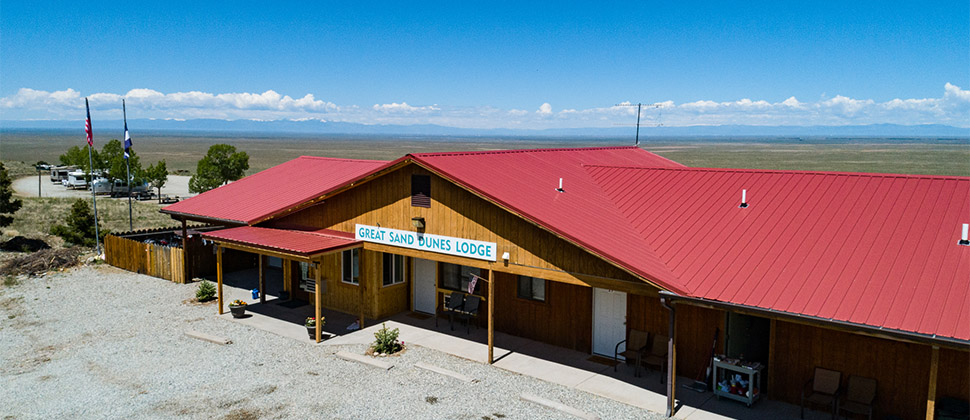 Exterior view of Lodge with signage over the door. Strands of clouds fill the background sky contrasted by the dark hues of mountains, and the gradient blues of the open sky. 