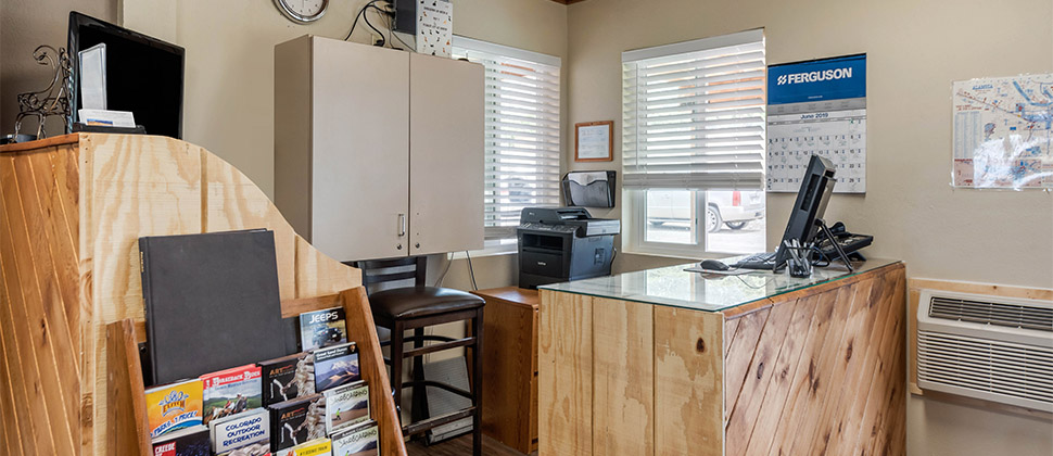 A small office/check-in area with glass countertop, pc and printer in the corner, and cabinets with a rack card display. 