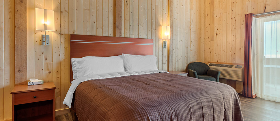 Brown bed and furniture compliment the soft bare wood walls. Lamps and side tables frame the bed with view of entry. 
