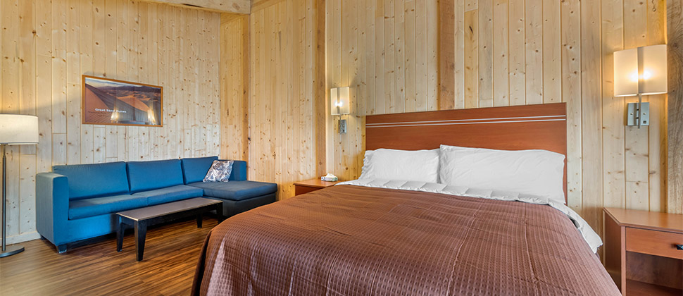 A bare wood room accented by brown bed and bed spread, and blue sofa in the corner. Lamps and artwork lightly grace the walls surrounding the bed.