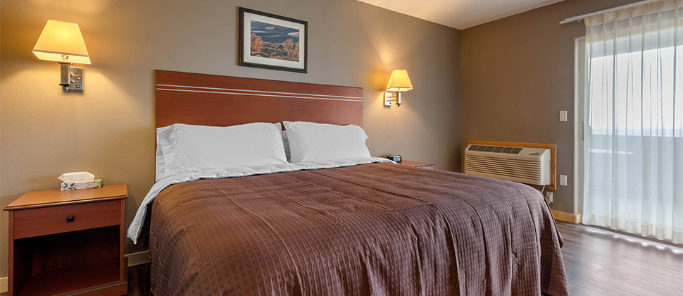 A bed centered between wall mounted lamps and matching bedside tables, with clear curtain over exterior doorway in the background. 