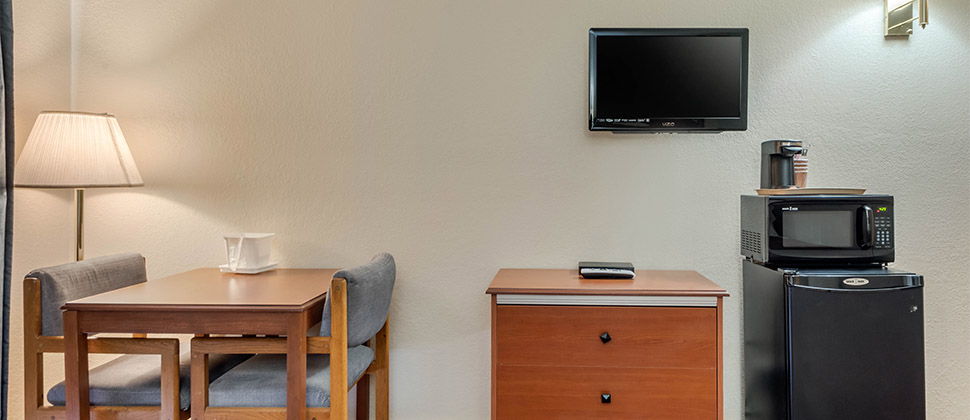 A television on the wall above a dresser is flanked by a small sitting area and a microwave atop a mini-fridge. 