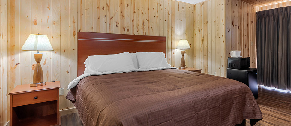 A bed situated between two bed side tables with matching lamps, and alcove with fridge and exterior window with charcoal drapes.