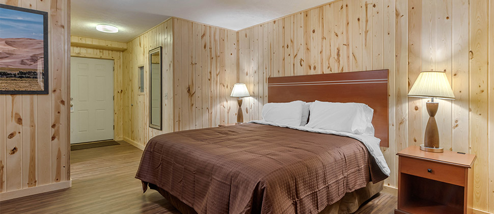 A bare wood room accented by brown bed and bed spread, and blue sofa in the corner. Lamps and artwork are mounted on the walls surrounding the bed.The entry hall and mirror can be seen in the background.