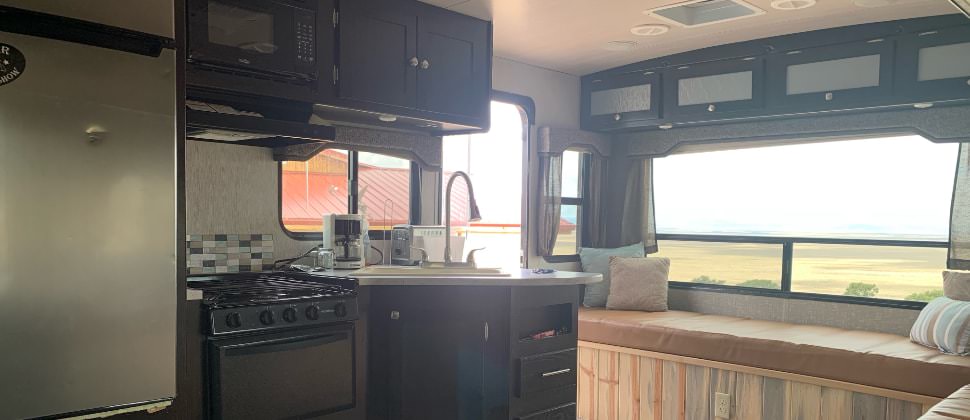 View of kitchen in camper with stainless steel refrigerator, black stove and microwave, dark wooden cabinets, and leather furniture